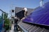 Zwei Männer installieren Solarmodule auf einem dunklen Ziegeldach, mit einem Baugerüst links und weiteren Modulen rechts.