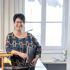 Lächelnde Frau mit Brille, kurzen dunklen Haaren und gemusterter Bluse hält eine schwarze Mappe im Büro.