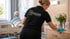 Pflegerin im schwarzen "SCHULDIS" T-Shirt reinigt eine Kommode in einem Krankenhauszimmer mit Patient im Bett.