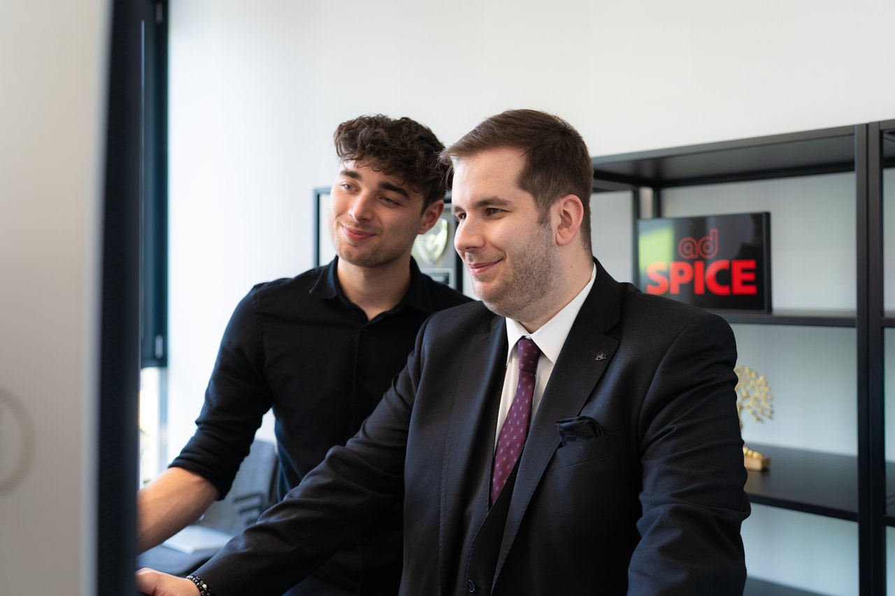 Drei Personen, zwei Männer und eine Frau, sitzen an einem Holztisch und schauen auf einen Laptop.