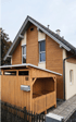 Kleines Haus mit brauner Holzfassade, Schrägdach, zwei Fenstern und einem Holzanbau mit Briefkasten und Hausnummer 123.