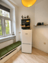 Weißer Kühlschrank in einer hellen Küche neben einer Sitzbank mit grünen Polstern und einem Fenster.