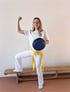 Lächelnde Frau in weißem Karate-Gi mit gelbem Gürtel und Pratzen, ein Bein auf einer Holzbank.