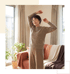 A smiling woman in a brown knit sweater and pants stretches her arms up in a sunlit living room with plants and a sofa.