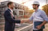 Zwei Männer, ein Geschäftsmann und ein Bauarbeiter mit Helm, schütteln sich die Hände auf einer Baustelle.