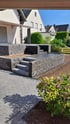 Neu gebaute Gartenterrasse und Treppenstufen aus dunkelgrauen Mauersteinen vor einem Einfamilienhaus.