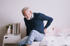An older man sitting on a bed, holding his back.