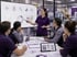 Woman in purple polo presenting data visualizations on a screen to four colleagues in a factory setting.