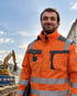 Ein lächelnder Mann in einer orangefarbenen Warnschutzjacke steht vor einer Baustelle mit einem gelben Bagger.