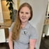 Junge Frau mit langen blonden Haaren und grauem Poloshirt mit "Fitness Für Frauen" Logo.