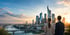 Zwei Personen blicken von hinten auf die Skyline von Frankfurt am Main mit Hochhäusern, Fluss und Brücken bei Sonnenuntergang.
