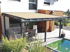 Moderne Terrassenüberdachung mit beigem Sonnenschutz, darunter Esstisch, Sofa und Pool.