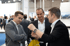 Drei Männer in Anzügen unterhalten sich auf einer Messe, einer zeigt auf seine Uhr.