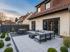 Moderne Terrasse mit grauen Fliesen, dunklem Gartenmöbel-Set, Feuerstelle und Zierpflanzen vor einem Haus.