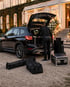 Ein Mann lädt schwarze Audiokoffer und Taschen in den offenen Kofferraum eines schwarzen BMW SUV vor einem Herrenhaus.