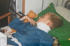 Ein blondes Kind mit Halskrause liegt auf dem Rücken im Bett, ein Teddybär liegt auf seiner Brust.