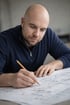 Ein Mann mit Glatze und Bart in einem dunkelblauen Hemd zeichnet mit einem Bleistift auf Bauplänen.