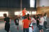 Ein Trainer im orangefarbenen T-Shirt leitet eine Gruppe Kinder beim Sportunterricht in einer Turnhalle an.