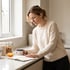 A woman in a cream sweater leans on a kitchen counter reading a notebook next to a mug and jars of cereal.