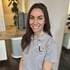 Eine lächelnde Frau mit langen braunen Haaren und einem grauen Poloshirt mit der Aufschrift "Fitness für Frauen".