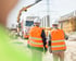 Zwei Ingenieure in weißen Schutzhelmen und orangefarbenen Warnwesten gehen auf einer Baustelle mit einem Lastwagen.