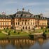 Barockes Schloss Pillnitz mit gelber Fassade, grünem Garten und Wassergraben unter blauem Himmel.