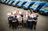 Gruppe von 26 Männern in Arbeitskleidung vor einer Reihe blauer Lieferwagen.