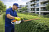 Mann in blauer Uniform und gelben Handschuhen schneidet eine Hecke mit einer weißen STIHL Heckenschere vor modernen Gebäuden.