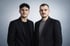 Zwei junge Männer in schwarzen Sakkos stehen vor einem hellgrauen Hintergrund.