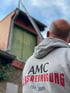 Glattköpfiger Mann von hinten mit grauem Kapuzenpullover "AMC GLASREINIGUNG EST. 2005" vor einem Haus mit Leiter.
