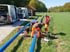 Drei Arbeiter in orangefarbenen Hosen verlegen blaue Rohre auf einer grünen Wiese neben zwei weißen Transportern.