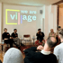 Vier Personen sitzen auf einer Bühne vor einer Leinwand mit dem Text "we are village", Publikum im Vordergrund.