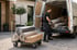 Ein Mann lädt Teppichrollen und Kartons aus einem weißen Lieferwagen, während ein Rollwagen mit weiteren Teppichen daneben steht.