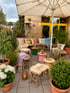 Eine gemütliche Terrasse mit Holzbänken, Kissen, Pflanzen in Töpfen, Hockern und einem großen Sonnenschirm.