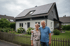 Glückliches Paar vor einem Haus mit Solarmodulen auf dem Dach und einem Holzzaun im Vordergrund.