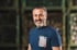 Lächelnder Mann mit grau meliertem Bart und dunkelblauem T-Shirt vor unscharfem Hintergrund in einem Lager.