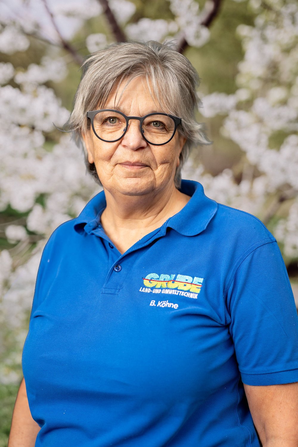 Frau mit grauen Haaren und Brille in blauem Poloshirt mit "GRÜN" Logo vor weiß blühendem Baum.