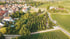 Luftaufnahme eines Dorfes mit Häusern, Bäumen, Weinbergen und einem Friedhof am Straßenrand.