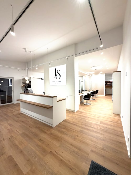 Heller Friseursalon mit Holzboden, weißer Rezeption und schwarzem JS-Logo an der Wand.