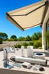 Beige Markise mit Sensoren, Fernbedienung und Montagezubehör auf einer Terrasse vor grünem Garten.