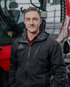 Ein Mann in schwarzer Jacke mit Logo und rotem Hemd lächelt vor einem roten Fahrzeug.