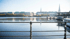 Blick über die Alster in Hamburg mit Gebäuden, Bäumen und einem Geländer im Vordergrund.