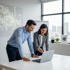 Two smiling business professionals, a man and a woman, look at a laptop screen in a bright office.