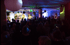 Live-Band auf der Bühne mit Instrumenten vor einem Publikum in einem dunklen Raum mit bunten Lichtern.