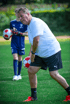 Ein erwachsener Mann hält einen Fußball, während ein Jugendlicher im Hintergrund einen weiteren Fußball hält.