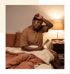 A woman sits on a bed holding a hand fan and resting her arm on her forehead next to a lit bedside lamp.