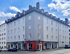 Eckgebäude mit grauer Fassade, roten Akzenten im Erdgeschoss und vielen Fenstern unter bewölktem Himmel.