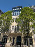Mehrstöckiges Wohn- und Geschäftshaus mit hellgelber Fassade, blauen Fensterrahmen im Obergeschoss und grünen Bäumen davor.
