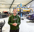 Mann im grünen Hoodie mit "5inline"-Logo steht in einer Autowerkstatt mit Rennwagen auf Hebebühne.
