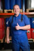 Lächelnder Mann in blauer Arbeitskleidung mit "BEST Chemie" Logo, Daumen hoch, vor Regalen mit blauen Fässern.